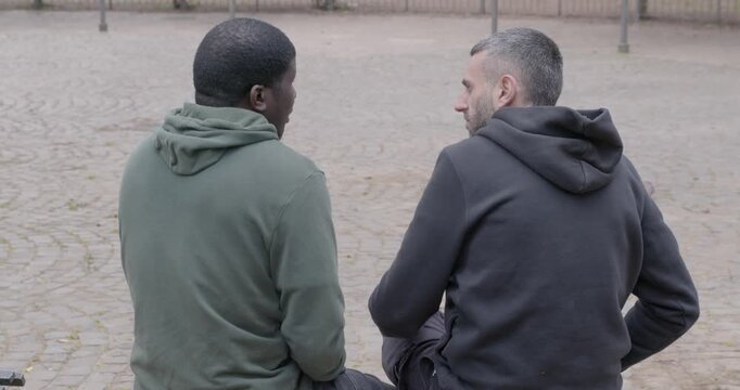 Two friends in hoodies talk about delicate personal issues in a park. Seen from behind, one comforts the other with a hug, expressing empathy, trust and emotional connection.