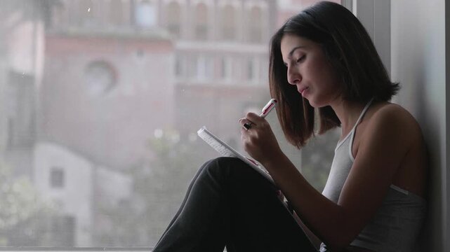 Dark-haired woman writes in her diary reflecting on recent problems. The scene conveys introspection, emotional release, and personal storytelling in a quiet moment.