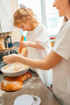 Caucasian Mother Teaching Daughter To Whisk Batter, Flourdusted Bowl On Wooden Countertop, Measuring Cups