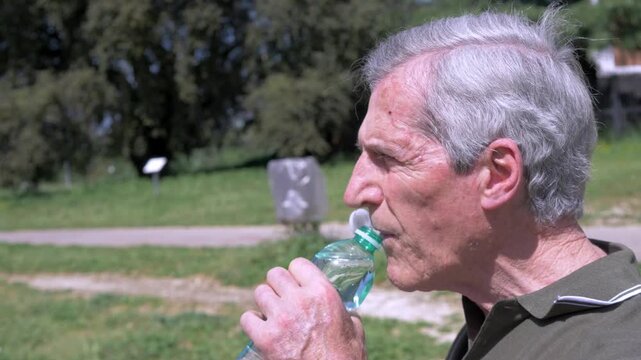 Side view of an elderly man drinking water from a bottle on a park bench during extreme summer heat, highlighting dehydration risk and the importance of hydration for health.