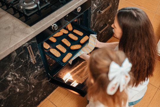 Mother And Daughter Baking Cookies Together, Pulling Tray From Oven In Modern Marble Kitchen, Mother Wearing