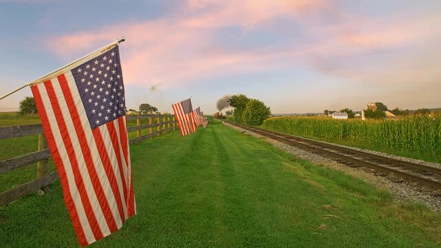 A train moves along the track beside tall grass. American flags wave gently in the breeze. The sun sets in the background, painting the sky with warm colors. This space shows simple rural life.