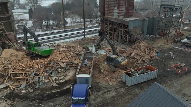 Workers use heavy equipment to remove wood debris from a construction site in winter. Trucks are parked nearby, and tracks can be seen in the background. The area is active with machines in use.