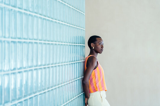 Focused Black woman leaning on glass block wall, looking aside with calm thoughtful emotion, creative professional pause, modern minimalist interior, urban lifestyle, copy space