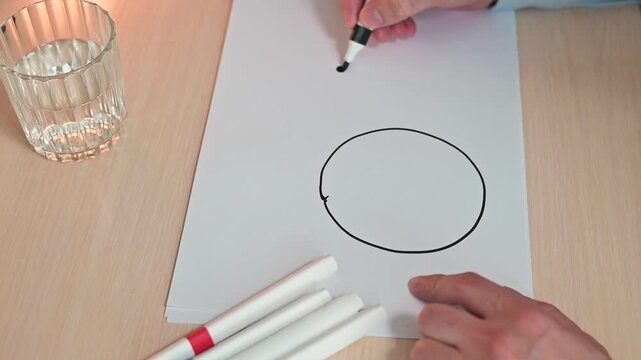 A hand uses a black marker to draw a circle on paper. The movement is intentional and precise, showing the process of marking a shape in a controlled setting
