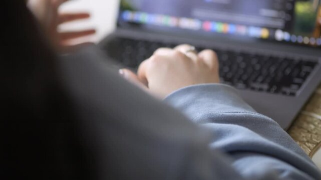 Over-the-shoulder view of person typing on laptop. Hands visible, fingers moving across keyboard. One hand holds a white cup near screen. Laptop screen shows colorful interface elements