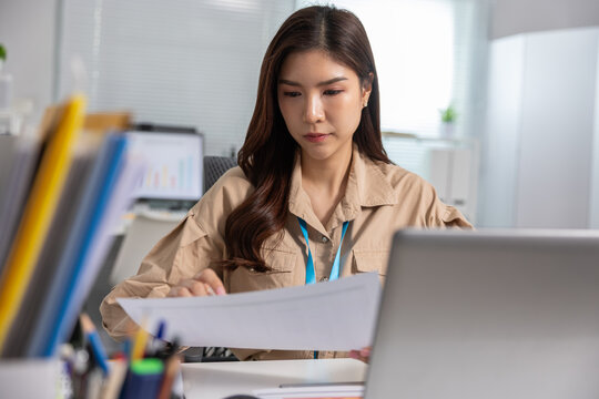 Serious young Asian business woman working at office desk reading document. Professional focused employee reviewing paper report near laptop computer showing intense concentration