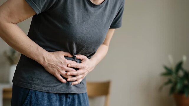 Person holding stomach in pain wearing casual t shirt indoors showing discomfort ache cramps abdominal pain digestive problem health issue hand suffering female in home setting