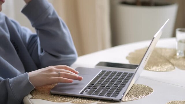 Person uses laptop on round table with gold patterned placemats. Hand rests near keyboard, fingers slightly curled. Person wears blue hoodie, elbow on table. Background features soft lighting