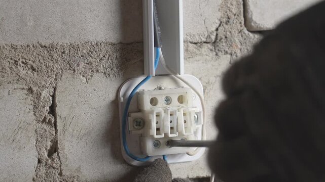 Close-up view of a professional electrician wearing protective gloves while using a screwdriver to connect wires in a terminal block on a rough brick wall during an electrical installation