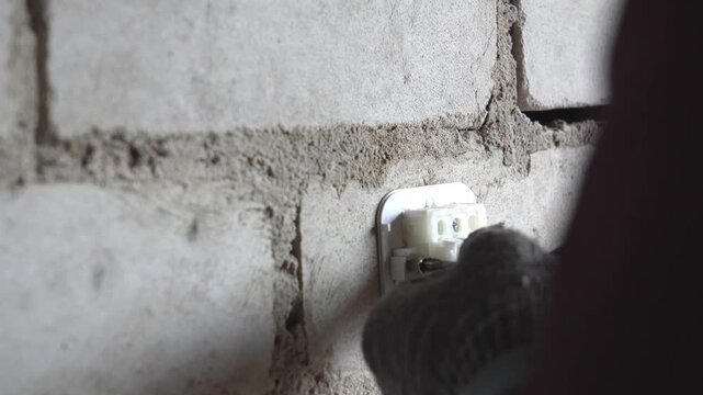 Electrician's hand in glove using a screwdriver to install a plastic switch and terminal block on an unfinished white brick wall during home renovation or construction