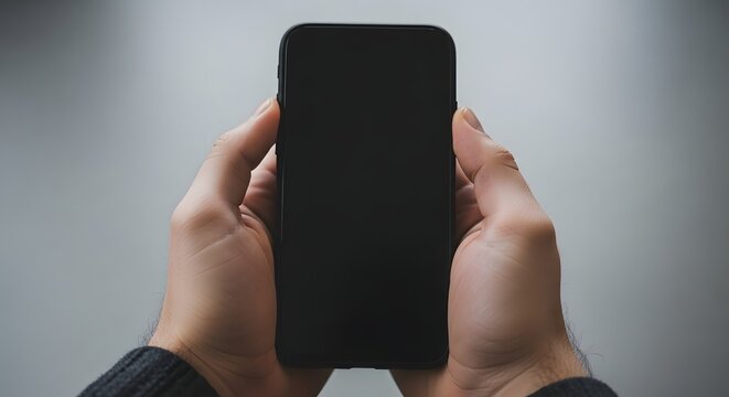A person holding a black smartphone with both hands in front of a gray background, closeup view of mobile phone