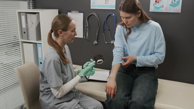 Female dermatologist wearing grey scrubs and medical gloves showing dermatoscope to young patient dressed in blue shirt and jeans, and examining her skin in clinic