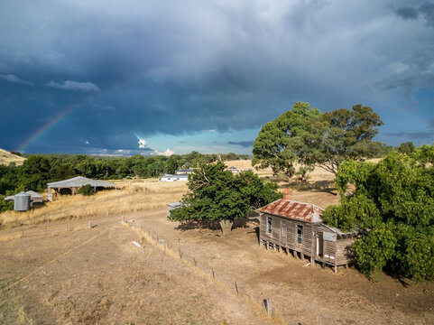 Aerial view of an abandoned farmhouse in a dry paddock under a dramatic sky and faint rainbow