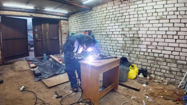 Industrial worker wearing a protective mask welding metal parts in a messy garage, with bright sparks flying from the electrode during the fabrication process in the workshop
