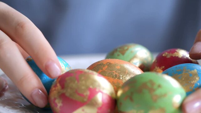 Hands gently pick colorful, gold-embellished Easter eggs. Fingers carefully handle decorated eggs. Background softly blurred with shallow depth of field. Egg colors include blue, green, red