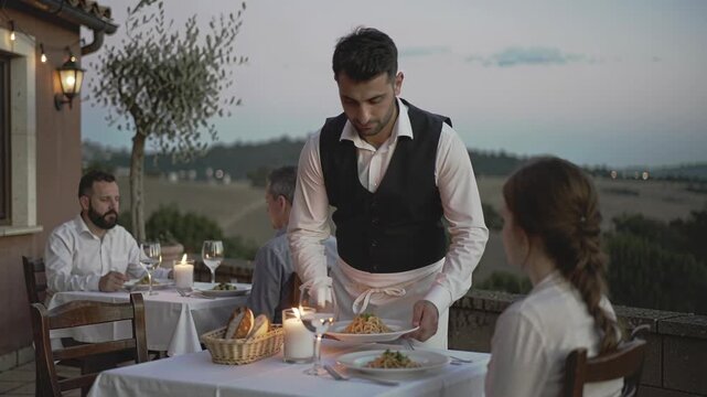 Man waiter serving dinner to a woman and men at an outdoor restaurant during evening. Dining experience with table service on a patio with a view.