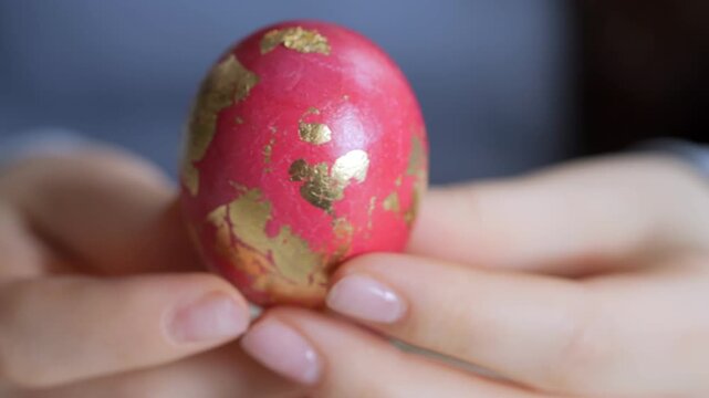 Hands gently hold a red egg with gold leaf patterns. Focus shifts from blurry background to detailed egg surface. Soft lighting highlights delicate gold accents on eggshell