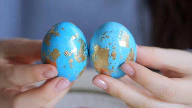 Hands hold two blue eggs with gold patterns. Eggs resemble decorative art or craft project. No animals or plants visible. Soft focus background, close-up shot. Detailed textures on egg surfaces