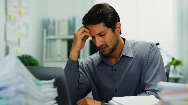 Tired man at desk, upset over project deadlines and paperwork stacks, showing burnout and mental fatigue.