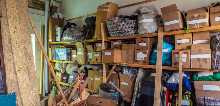 A cluttered, messy wooden shelf in a storage area is filled with disorganized cardboard boxes, plastic bags, plaid storage bags, and various loose items, reflecting a busy or untidy environment.