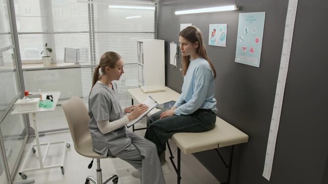 Young female gynecologist filling medical form, then gen Z patient standing up from couch and coming to grey wall, and doctor measuring her height