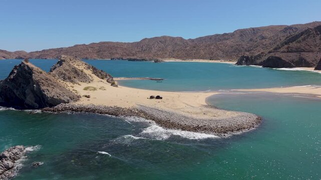 An aerial view shows an SUV and teardrop camper parked on a beach island at Punta Final in Baja California during a sunny day.