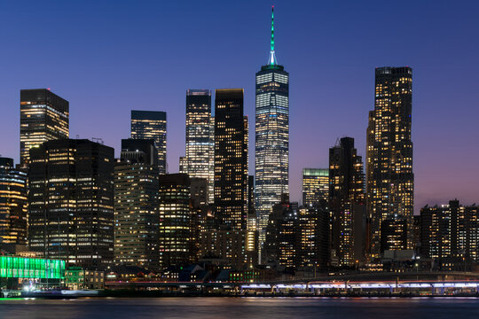 Skyscrapers with glowing windows stand densely packed in downtown Manhattan as seen from across the East River at night.
