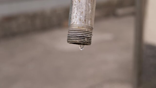 A close-up view of an old rusty metal pipe with threads dripping water slowly against a blurred background. Concept of water waste, plumbing maintenance, drought, and resource conservation.