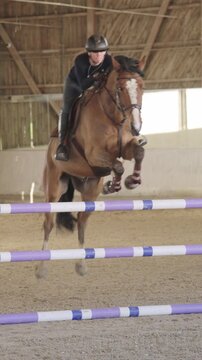 Young female equestrian practicing show jumping on her brown horse in a large indoor riding arena
