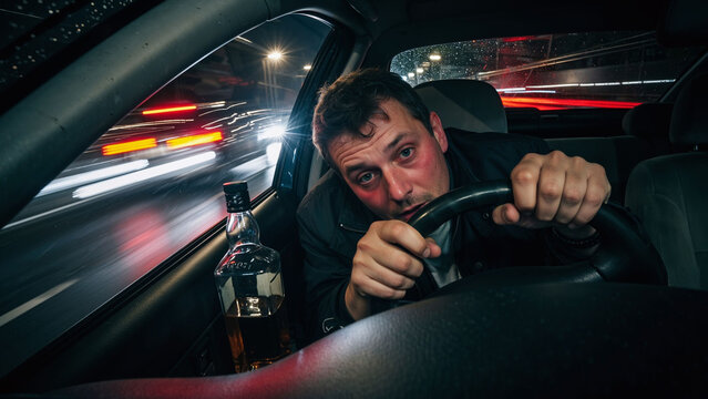 Man with red face driving car at night with bottle of alcohol beside him. Concept of drunk driving. Social problem.