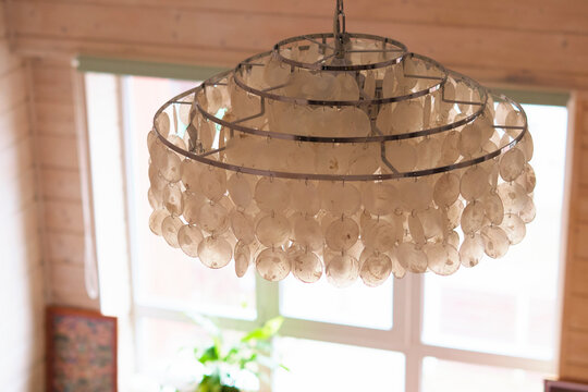 Close up of a shell chandelier hanging in front of a bright window inside a warm wooden room. Natural textures and soft daylight bring boho style, home comfort, and relaxed interior design.