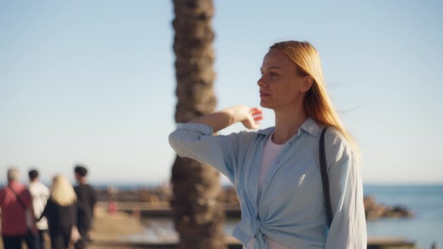 Blonde woman enjoying warm sunny weather on seafront promenade, pausing to feel the sea breeze and light. Relaxed vacation moment, coastal lifestyle, travel escape, summer atmosphere.