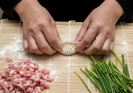 Top down view of weathered hands expertly pleating a fresh dumpling on a bamboo mat with raw minced pork and fresh green chives arranged on the table.
