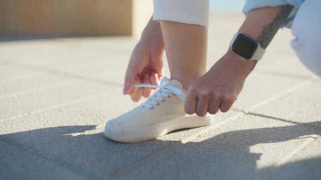 Close-up of woman tying shoelaces on white sneakers while walking outdoors on sunny day. Everyday lifestyle moment on city sidewalk, casual preparation, urban travel and walking concept.