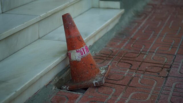Neglected urban environment with weathered orange traffic cone covering a hole from missing paving stone on red sidewalk next to marble steps. Urban decay, infrastructure damage, safety hazard concept
