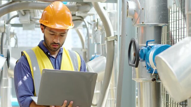 Professional engineer in hard hat and safety vest uses a laptop to monitor industrial pipe systems and adjust valves in a factory plant. Maintenance, technology, and engineering service concept.