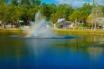 Fototapeta premium Neighborhood Lake Park Fountain With Rainbow Spray, Palm Trees and Waterfront Houses on Sunny Day in Gulfport, FL. Creating a vivid rainbow spray, palm trees lining the shore and pastel waterfront hou