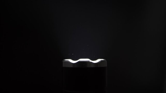 Ashtray sits on a black background as smoke and ash particles rise. Dramatic top lighting creates high contrast against the dark, isolated background.