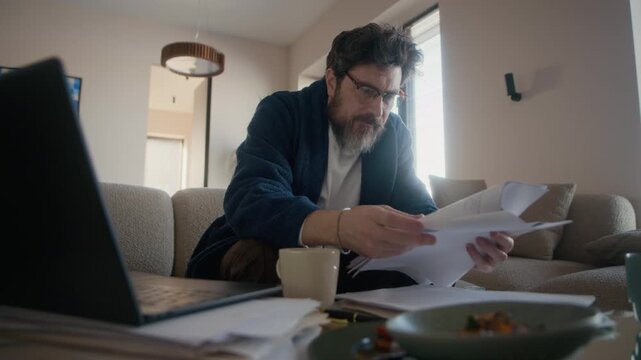 Medium shot of mature Caucasian writer with beard on face and messy hair wearing robe and eyeglasses sipping coffee while working on new novel