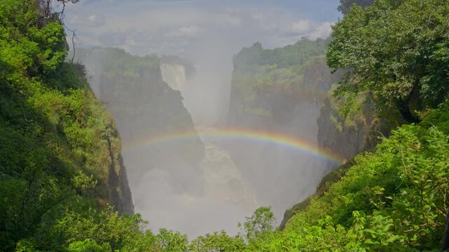 Mosi oa Tunya, aka Victoria Falls, shows immense power with water cascading down, creating mist and vibrant rainbow spanning canyon on Zambezi River
