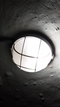 Industrial bulkhead light with a protective cage flickers on a rough concrete ceiling. The erratic illumination creates a harsh, eerie glow in a dark, suspenseful environment.