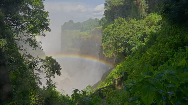 People watching Victoria Falls with magnificent rainbow forming in mist on Zambezi River at border between Zambia and Zimbabwe, majestic natural wonder in Africa