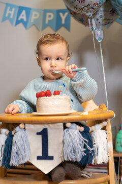 Toddler Baby boy eating himself birthday cake by spoon. First birthday family celebration. Child celebrates birthday with cake and decorations at home with balloons and decoration. Vertical card.