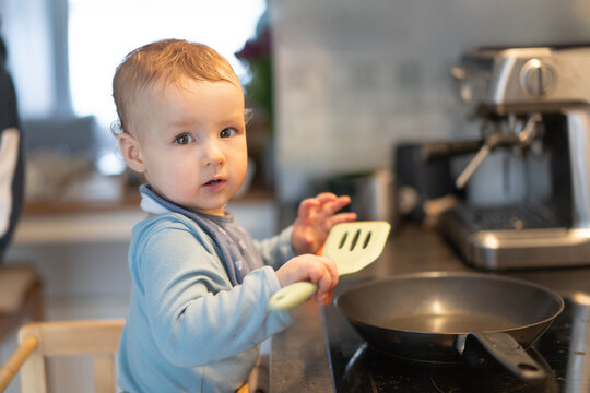 Baby boy standing on learning tower near induction stove with frying pan and holding a spatula in hand. Toddler repeating actions after parents, learning to cook food. Everyday kid development routine