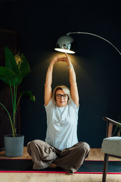 Middle aged woman on floor mat and doing stretching yoga position in modern Scandi dark interior home. Healthy Lifestyle. Sport activity for mature wellbeing. Mindful movement, relaxation. Vertical.