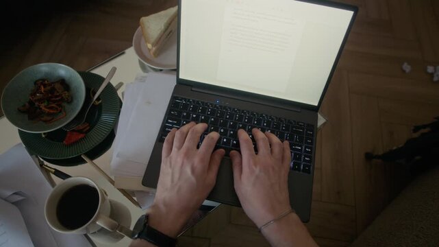 High angle POV of hands of unrecognizable male author writing novel on laptop and drinking coffee, food, plates with leftovers and sandwich, paper drafts visible on coffee table