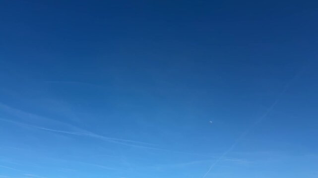 Sky showing a clear blue expanse with visible airplane contrail, a distant moon appearing in bright daylight, representing vast atmospheric space, environmental calm.
