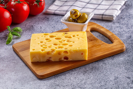 Block of Swiss cheese on a wooden cutting board with grilled olives and fresh tomatoes