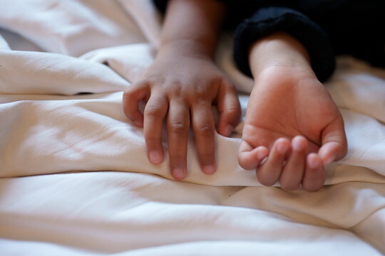Close-up of a young child's hands resting peacefully on a white textured bed sheet. The soft lighting emphasizes the gentle skin and small fingers of the little girl during a moment of rest.